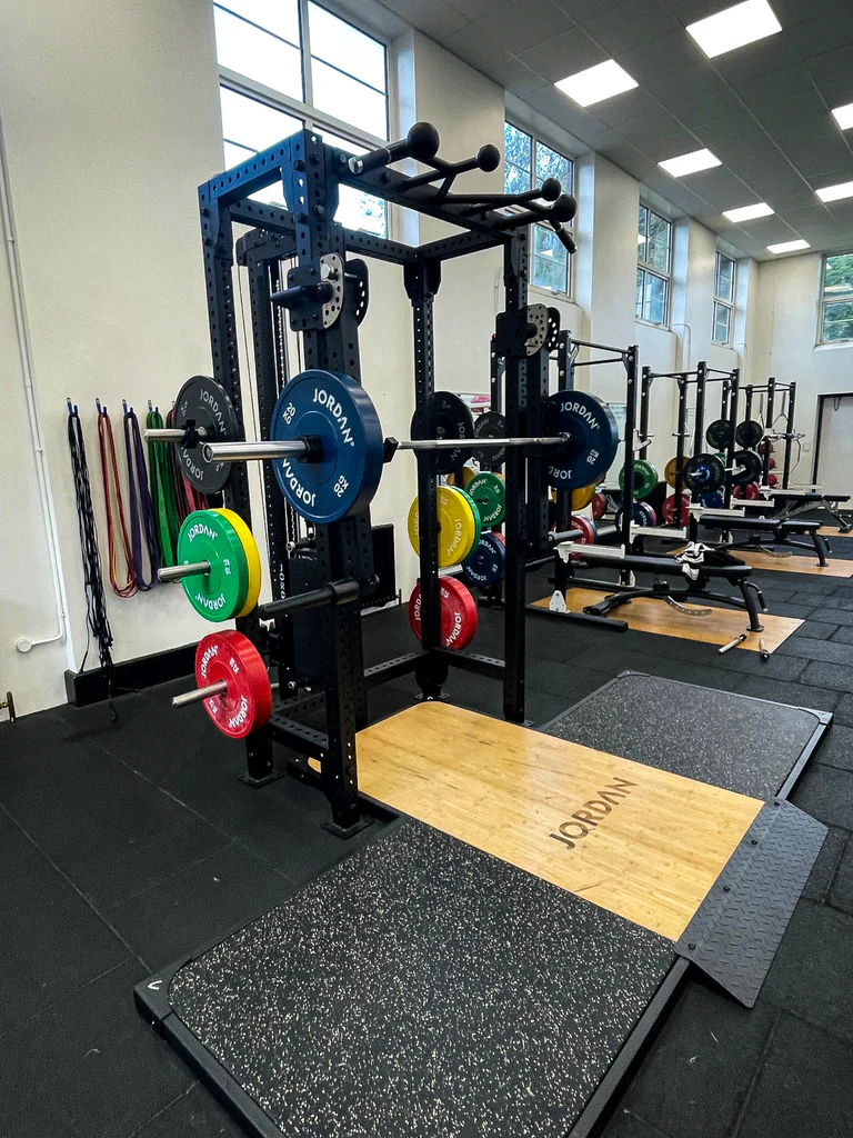  JORDAN® J75 T-Rack Displaying J Hooks, Cable and Pull up bar, displaying weight plates on the storage pegs and barbell on j hooks in a gym setting 